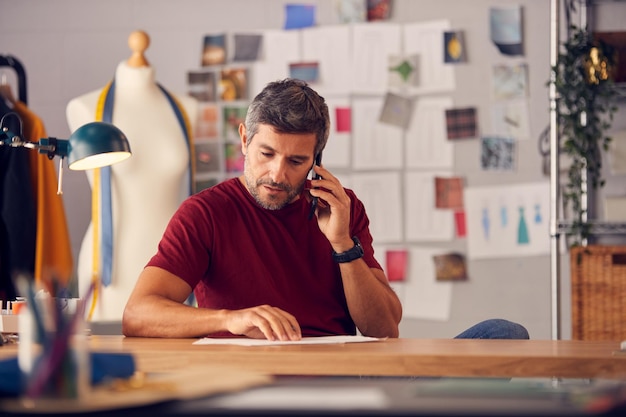 Créateur de mode masculin mature en studio travaillant sur des croquis ou des documents au bureau faisant un appel téléphonique