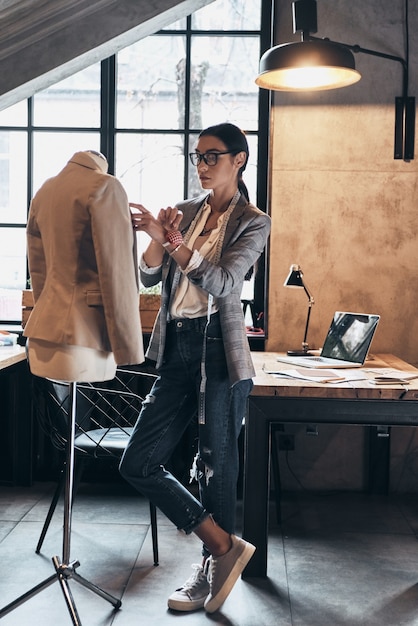 Créateur de mode à succès. Toute la longueur d'une jeune femme sérieuse en lunettes à l'aide d'aiguilles à coudre pour coudre une veste sur mannequin en se tenant debout dans son atelier