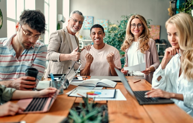 Des créatifs multiraciaux dans un bureau moderne. Un groupe de jeunes gens d'affaires et un patron senior travaillent avec un ordinateur portable, une tablette, un téléphone intelligent, un ordinateur portable, des graphiques. Une équipe performante en coworking