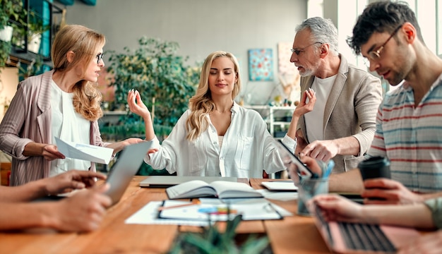 Des Créatifs Multiraciaux Dans Un Bureau Moderne. Un Groupe De Jeunes Gens D'affaires Et Un Patron Senior Travaillent Avec Un Ordinateur Portable, Une Tablette, Un Téléphone Intelligent, Un Ordinateur Portable, Des Graphiques. Une équipe Performante En Coworking