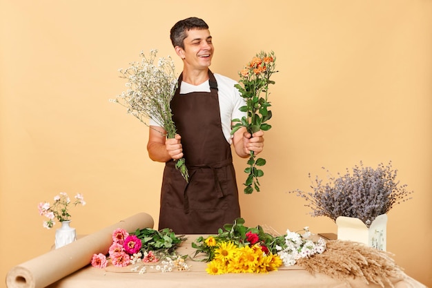Créations de bouquets colorés Propriétaire de magasin prospère Heureux jeune homme en tablier marron travaillant dans un magasin de fleurs debout isolé sur fond beige faisant une composition élégante