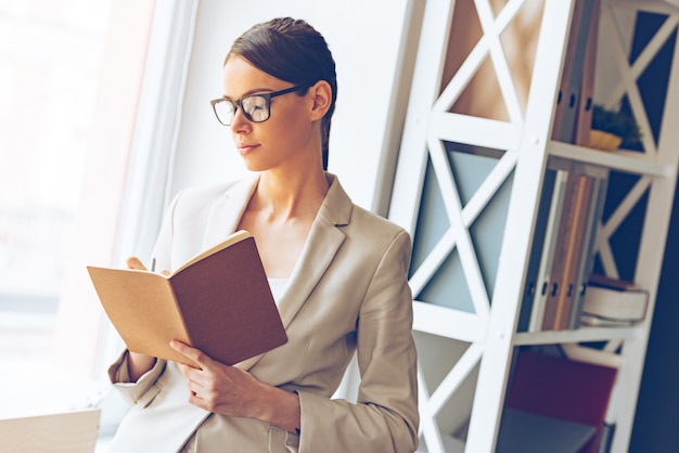 Créativité au travail. Jeune belle femme d'affaires dans des verres écrivant dans un ordinateur portable en se tenant debout sur le rebord de la fenêtre