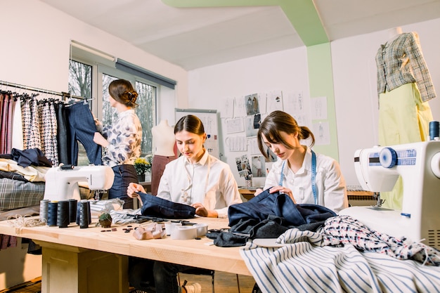 Créatrice de mode féminine debout près de porte-vêtements avec des vêtements à la main élégants à la mode. Deux jeunes filles couturières travaillant sur une nouvelle collection de pantalons pour les clients dans un atelier confortable