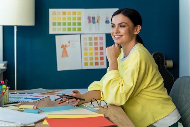 Créatrice de mode femme brune assise à la table et détournant les yeux avec un visage pensif en pensant à sa future collection de vêtements