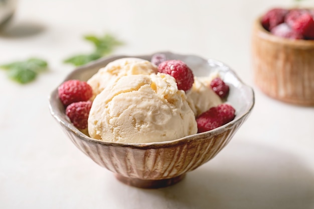 Crème glacée à la vanille caramel maison avec des framboises congelées dans un bol en céramique debout sur une table en marbre blanc avec des fleurs derrière.