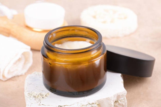 Crème hydratante ouverte dans un bocal en verre ambré sur podium en pierre et ensemble pour les produits de soins de la peau et du corps sur fond beige