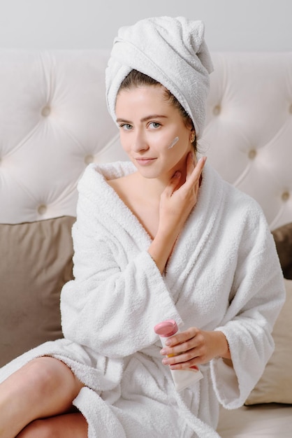 Crème pour le visage femme en peignoir avec une serviette sur la tête après une douche à la maison. appliquer de la crème pour la peau sur la peau.