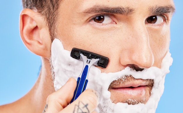 Photo crème de toilettage et homme se rasant le visage avec un rasoir pour les soins de la peau et la santé de bien-être sur un fond de studio bleu beauté jeune et portrait d'un modèle avec de la mousse sur la barbe pour l'hygiène