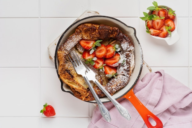 Crêpe de bébé hollandais avec baies de fraises fraîches et saupoudrées de sucre glace en poudre dans une poêle rouge sur fond de cuisine blanche Vue de dessus.