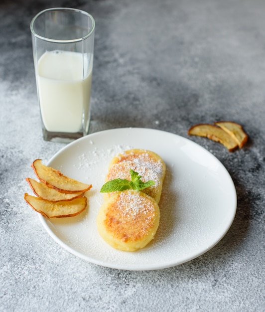 Crêpes au fromage cottage frais sur une assiette blanche avec un verre de lait sur un fond de béton. Petit-déjeuner sain et diététique