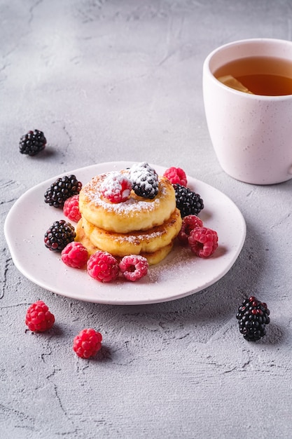 Crêpes Au Fromage Cottage Et Sucre En Poudre, Beignets De Caillé Dessert Avec Framboises Et Mûres Dans Une Assiette Près D'une Tasse De Thé Chaud Avec Une Tranche De Citron Sur Une Surface De Béton En Pierre, Vue D'angle