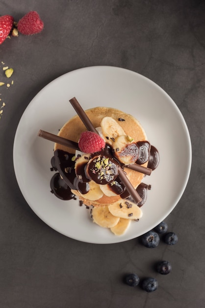 Crêpes aux fraises, framboises, banane, chocolat, pistaches et crème sur une plaque blanche.