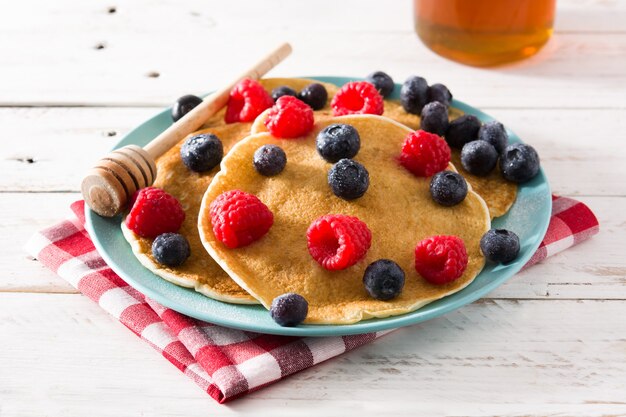 Crêpes aux framboises, bleuets et miel sur table en bois blanc.