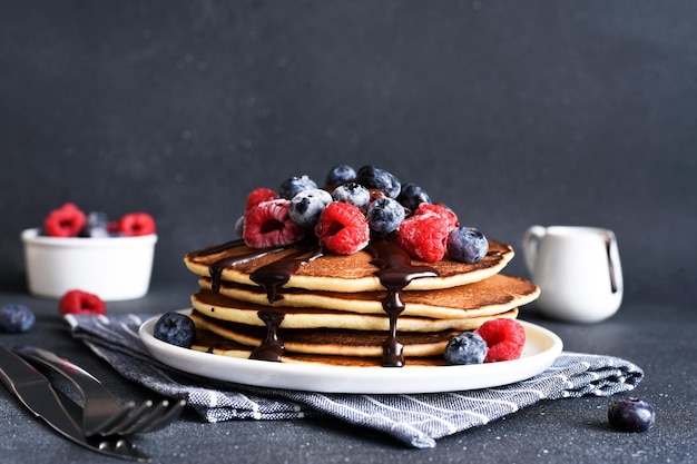 Crêpes aux framboises et bleuets et sauce au chocolat sur un fond de béton foncé Pile de crêpes aux baies