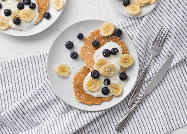 Crêpes à l'avoine faites maison avec du yaourt, des bleuets frais et de la banane à une table en bois blanc.