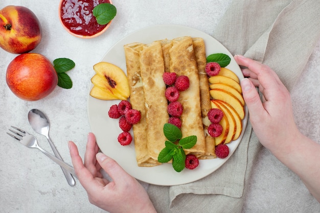 Crêpes, crêpes fines aux framboises et pêches, menthe et confiture sur une assiette avec des mains féminines