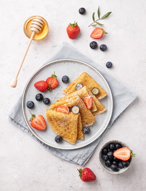 Photo crêpes ou crêpes fines faites maison à plat avec des myrtilles fraîches et des fraises et du miel sur une assiette sur fond clair vue de dessus