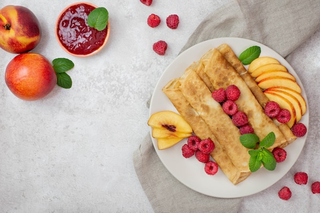 Crêpes, crêpes minces aux framboises, fruits de pêches, menthe et confiture de baies sur une surface gris clair