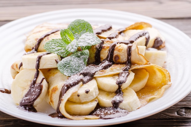 Crêpes françaises avec sauce au chocolat et banane dans un plat en céramique sur une table de cuisine en bois