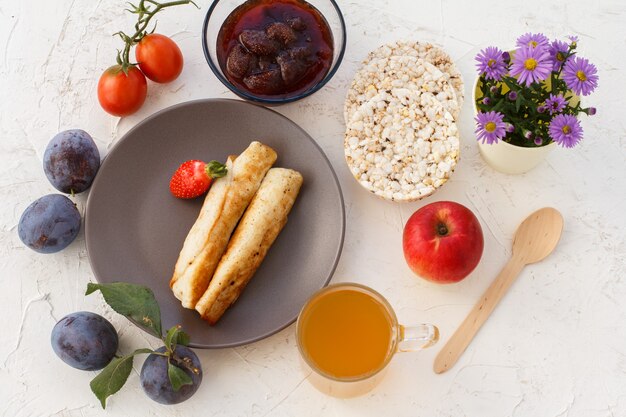 Crêpes maison fourrées au fromage cottage, une fraise dans l'assiette, des prunes, un bol en verre avec de la confiture, une cuillère en bois, des pommes, des galettes de riz soufflé, une tasse de thé et des fleurs. Vue de dessus.