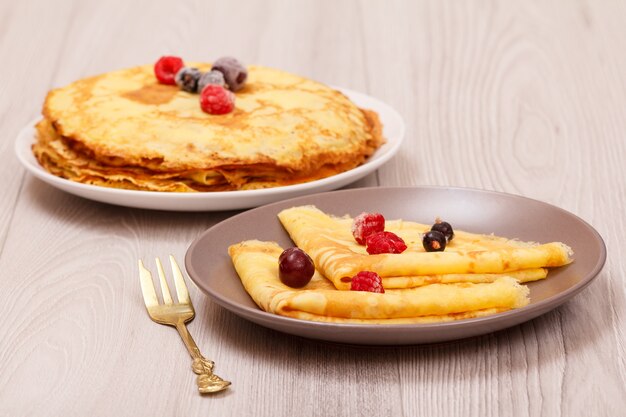 Photo crêpes maison garnies de framboises et de mûres congelées sur assiettes et fourchette en métal sur fond de bois.