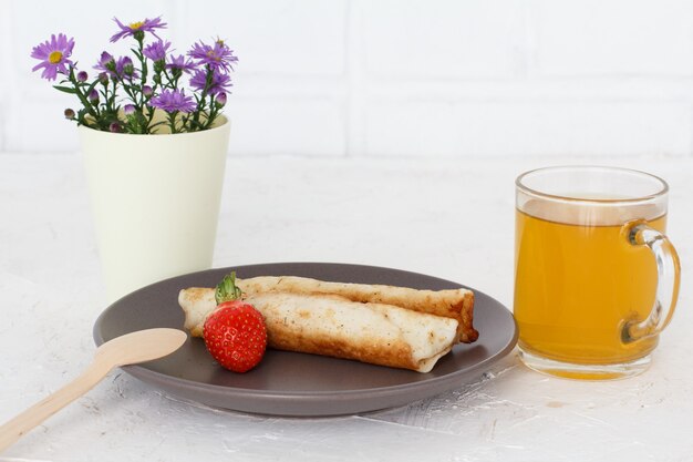 Crêpes maison remplies de fromage cottage, une fraise sur une assiette, une tasse de thé et des fleurs sur la surface blanche