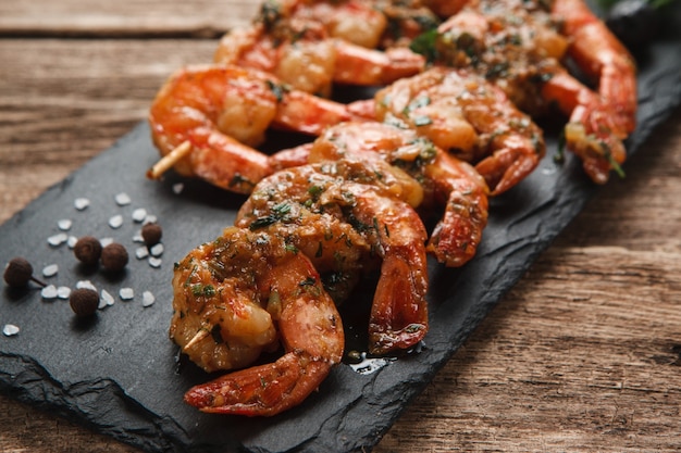 Crevettes épicées frites aux herbes sur des brochettes en bois servies sur une slade noire, vue rapprochée.