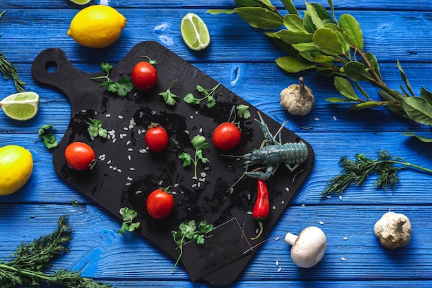 Écrevisses vivantes sur une planche à découper noire, sur un fond bleu de différentes couleurs à côté de légumes et de verts