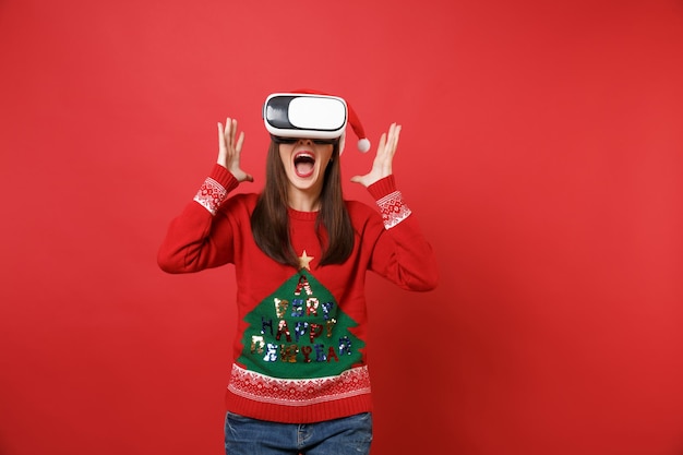 Crier la jeune fille de Santa en pull tricoté, chapeau de Noël regardant dans le casque, écartant les mains isolées sur fond rouge. Bonne année 2019 concept de fête de vacances célébration. Maquette de l'espace de copie.