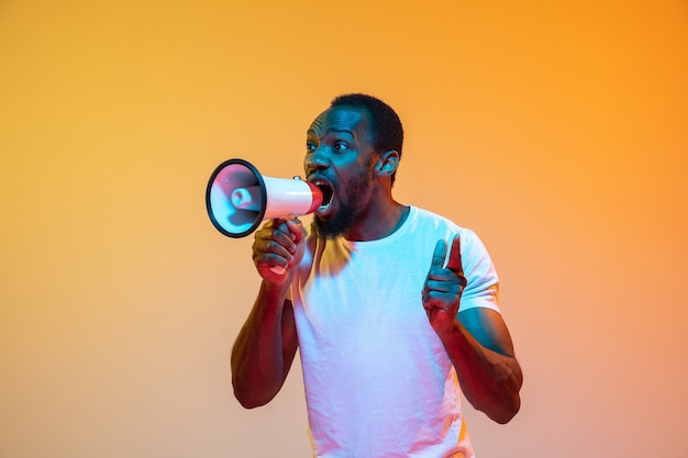 Crier avec mégaphone. Portrait moderne de l'homme afro-américain sur fond de studio orange dégradé au néon. Beau modèle afro. Concept d'émotions humaines, d'expression faciale, de ventes, d'annonces. Espace de copie.