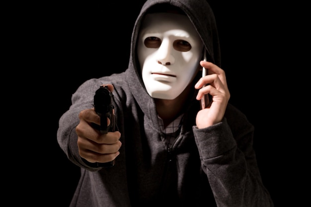 Photo criminel en masque utilisant un téléphone portable tout en ciblant avec un pistolet contre un fond noir