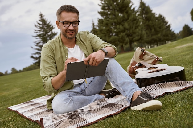 Écrire une chanson jeune homme créatif portant des lunettes assis sur une herbe verte dans le parc et faire