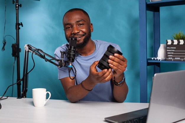 Un critique de technologie afro-américain souriant présentant les fonctionnalités de l'objectif zoom dslr dans un podcast en ligne en direct. Célèbre influenceur avec microphone examinant l'équipement de photographie devant un ordinateur portable.