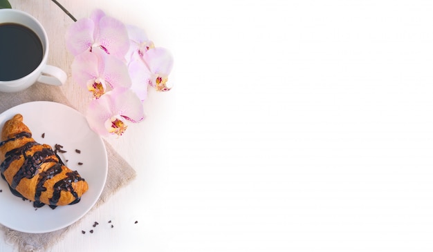 Croissant au chocolat, tasse de café et une orchidée rose sur fond blanc