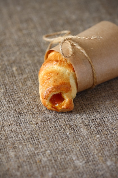 Croissant à La Confiture De Pêche Enveloppé Dans Du Papier D'emballage Kraft Et Attaché Avec Des Cordes Avec Des Arcs Sur Une Nappe De Toile De Jute Rugueuse.