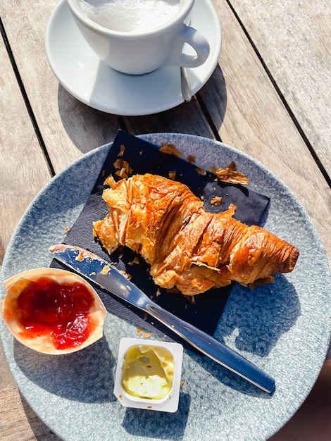 Croissant fraîchement cuit avec du beurre et de la confiture de baies et une tasse de café blanc sur une table en bois par une journée d'été ensoleillée. Vacances manger dans un café. Repas fini.