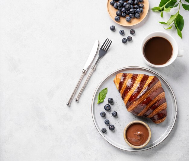 Croissant frais avec chocolat sur une assiette blanche sur un fond clair avec des bleuets et une tasse de café Délicieux concept de petit déjeuner fait maison