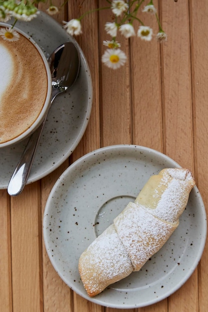 Croissant français fait maison avec de la poudre Tasse de cappuccino dans une tasse en céramique blanche avant-plan flou de fleurs de camomille sur une table en bois sur la terrasse Petit-déjeuner esthétique