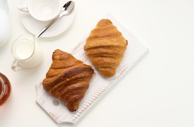 Croissants au four, théière en céramique blanche et tasse et soucoupe vides, pot de miel sur une table blanche, vue de dessus. Déjeuner