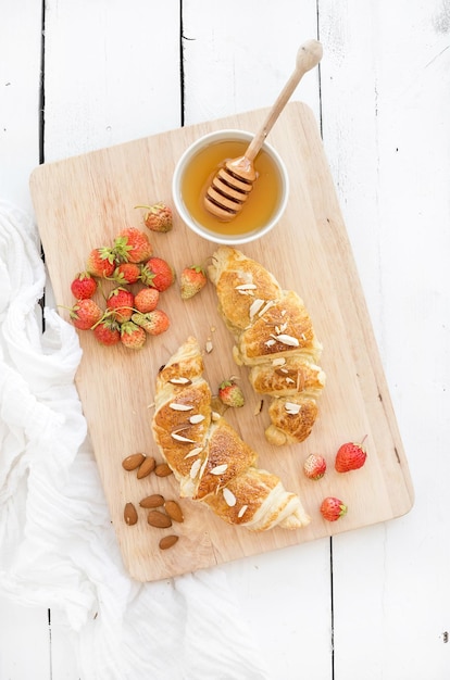 Croissants aux amandes fraîchement cuits au four avec fraises du jardin et miel sur planche de service sur fond de bois rustique blanc