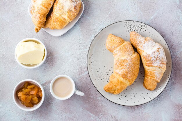 Croissants frais et croustillants sur des assiettes, une tasse de café et des bols avec de la confiture et du beurre sur un fond texturé clair. Pause café ou petit déjeuner. Vue de dessus