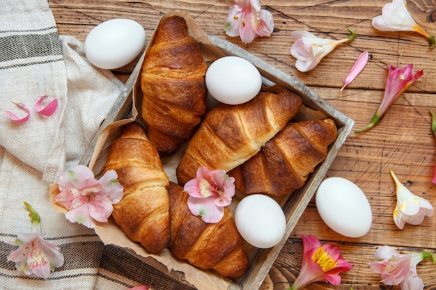 Croissants frais, œufs et fleurs dans un bac sur une table en bois vue de dessus