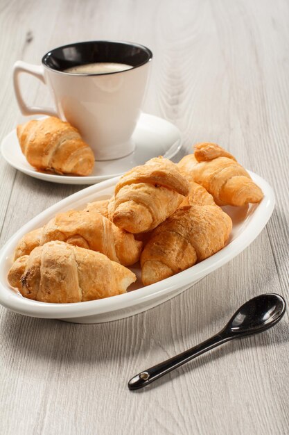 Croissants frais sur plat en porcelaine blanche, tasse de café noir avec soucoupe sur fond