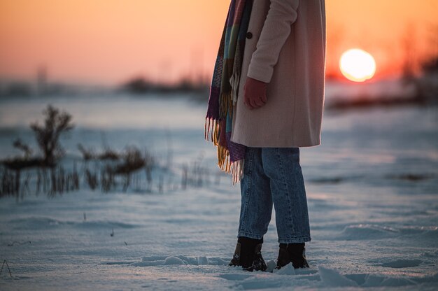 Crop femme méconnaissable dans des vêtements chauds et des bottes sur la neige blanche fraîche dans la campagne d'hiver au moment du coucher du soleil