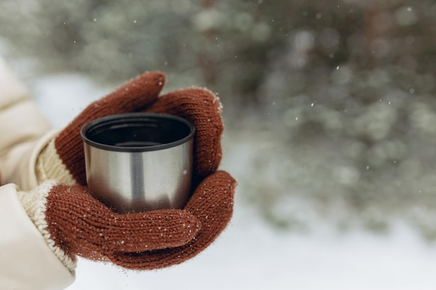 Crop femme en mitaines se réchauffant les mains avec du thé dans une tasse thermos dans les bois d'hiver enneigés