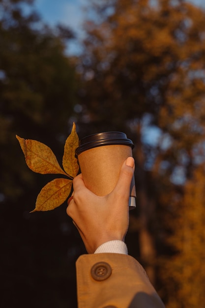 Crop femme avec une tasse de café pour aller avec des feuilles dans le parc d'automne au coucher du soleil