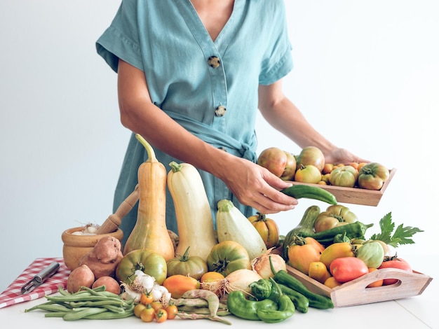 Crop inconnue femme en robe debout près de tas de citrouilles fraîches et de poivrons et d'oignons avec de l'ail et des pois placés sur la table et tenant des tomates contre le mur blanc