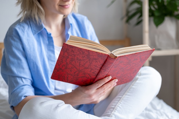 Cropped blonde woman holding open book et lecture assis sur le lit. Loisirs, séjour à la maison et mise en quarantaine.