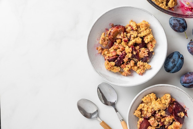 Crumble aux fruits. Tarte crumble aux prunes dans des assiettes avec des cuillères sur une table en marbre blanc pour un délicieux petit-déjeuner. Vue de dessus avec espace de copie