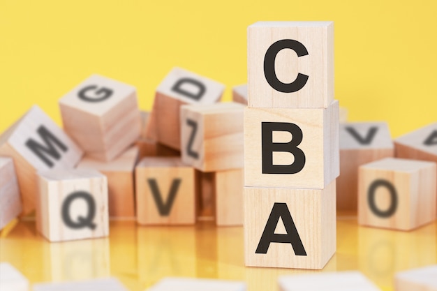Cubes en bois avec lettres CBA disposés en pyramide verticale, fond jaune, reflet de la surface de la table, concept d'entreprise. cba - abréviation de l'analyse coûts-avantages